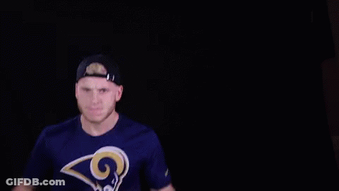 A Man Wearing A Rams T-Shirt And A Hat Is Holding His Hands Up In The Air . GIF