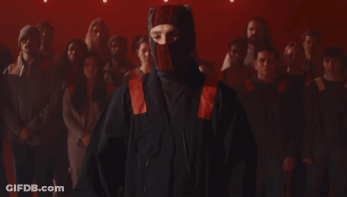 A Man Wearing A Red And Black Mask Stands In Front Of A Crowd Of People GIF
