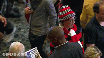 A Man Wearing A Red And Black Striped Hat Is Reading A Newspaper In A Crowd Of People GIF