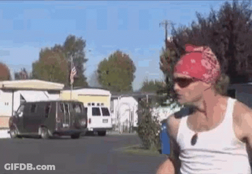 A Man Wearing A Red Bandana And Sunglasses Is Walking Down A Street In A Trailer Park . GIF