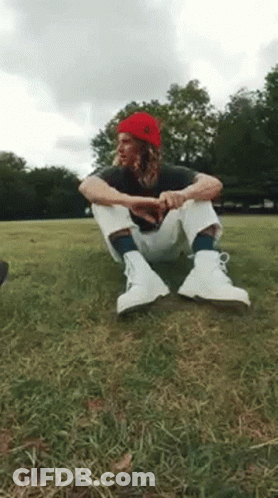 A Man Wearing A Red Beanie And White Boots Is Sitting On The Grass . GIF