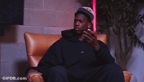 A Man Wearing A Red Bull Hat Sits In A Chair Looking At His Phone GIF