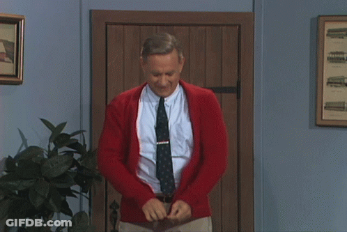 A Man Wearing A Red Cardigan And Tie Is Standing In Front Of A Wooden Door GIF