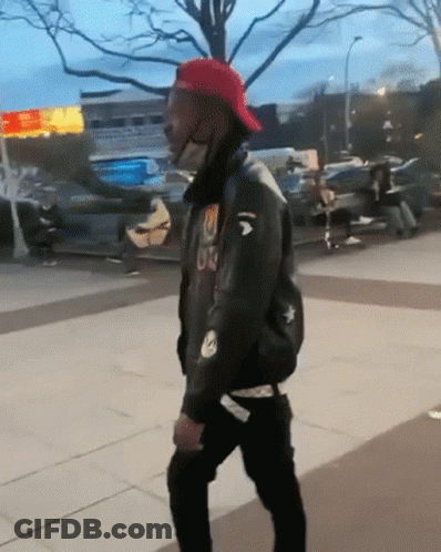 A Man Wearing A Red Hat And A Black Jacket With The Letter U On It Is Walking Down A Sidewalk GIF