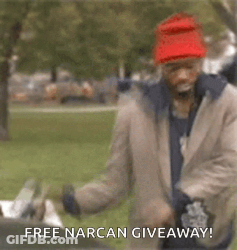 A Man Wearing A Red Hat And A Jacket Is Standing In A Park With The Words `` Free Narcan Giveaway ! '' GIF