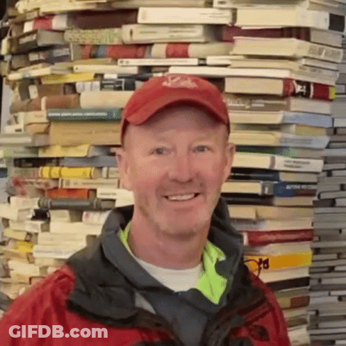 A Man Wearing A Red Hat And A Jacket Stands In Front Of A Pile Of Books GIF