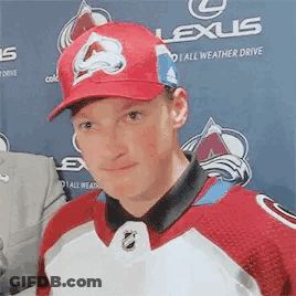 A Man Wearing A Red Hat And A Red And White Hockey Jersey Is Standing In Front Of A Lexus Banner . GIF