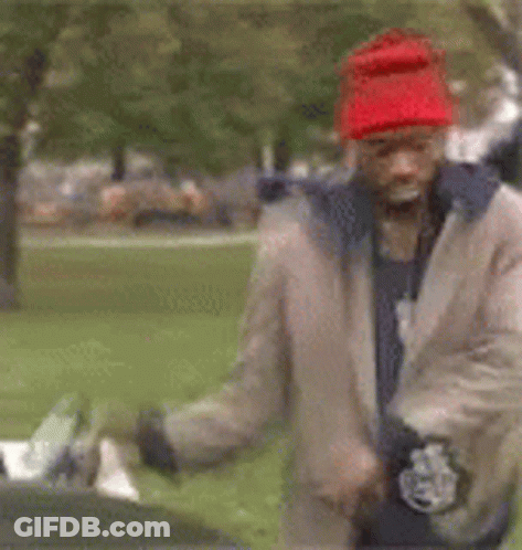 A Man Wearing A Red Hat And A Tan Jacket Is Standing In A Park . GIF