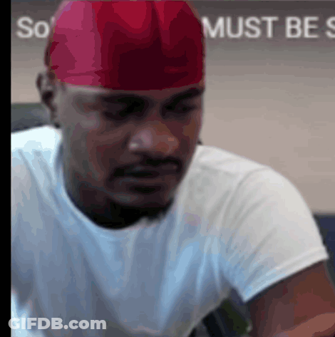 A Man Wearing A Red Hat And A White Shirt Is Sitting In Front Of A Sign That Says So Must Be So GIF