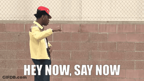 A Man Wearing A Red Hat And A Yellow Jacket Stands In Front Of A Brick Wall And Says Hey Now Say Now GIF