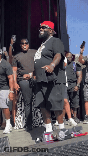A Man Wearing A Red Hat And Sunglasses Stands In Front Of A Crowd Of People GIF