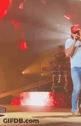 A Man Wearing A Red Hat That Says Supreme On It Is Singing Into A Microphone On A Stage GIF