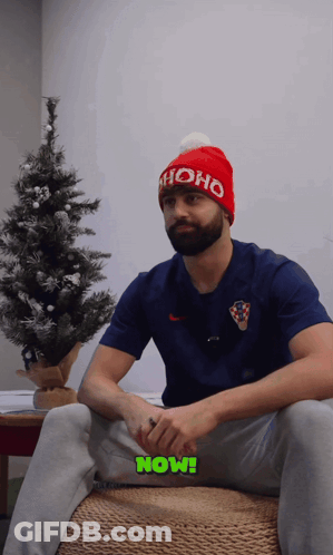 A Man Wearing A Red Ho Ho Hat Sits In Front Of A Small Christmas Tree GIF