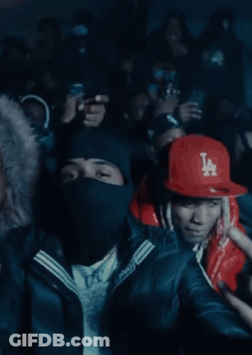 A Man Wearing A Red Jacket And A La Hat Stands In Front Of A Crowd GIF
