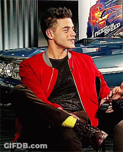 A Man Wearing A Red Jacket Sits In Front Of A Need For Speed Poster GIF