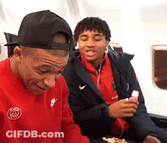 A Man Wearing A Red Nike Shirt Is Sitting Next To Another Man Wearing A Black Hat . GIF