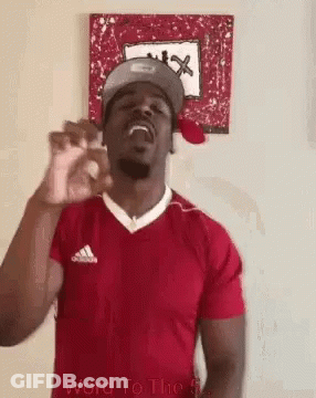 A Man Wearing A Red Shirt And A Hat Is Standing In Front Of A Painting . GIF