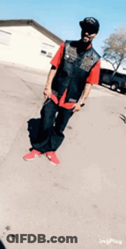 A Man Wearing A Red Shirt And Black Vest Is Standing On A Street GIF