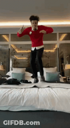 A Man Wearing A Red Sweater Is Jumping On A Bed GIF
