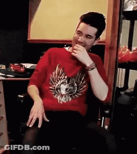 A Man Wearing A Red Sweater With A Wolf On It Is Sitting In A Chair And Laughing . GIF