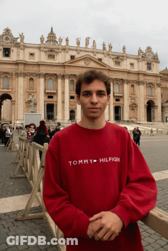 A Man Wearing A Red Tommy Hilfiger Shirt Stands In Front Of A Large Building GIF