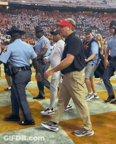 A Man Wearing A Red Visor Is Walking On A Field With Police Officers GIF