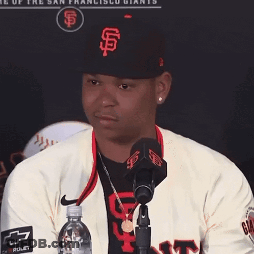 A Man Wearing A San Francisco Giants Hat Stands In Front Of A Microphone Holding A Bottle Of Water GIF