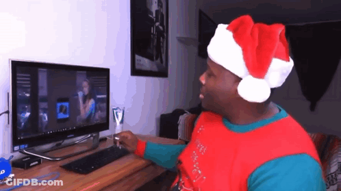 A Man Wearing A Santa Hat Is Sitting In Front Of A Computer Monitor GIF