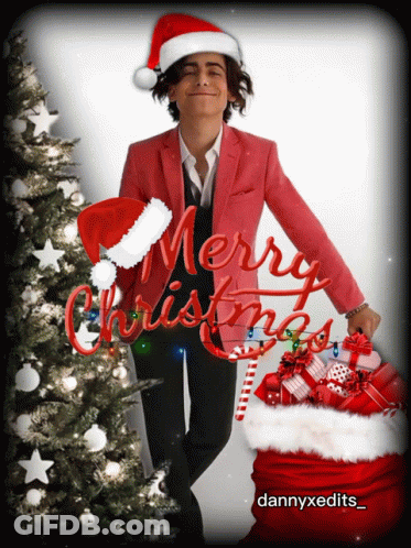 A Man Wearing A Santa Hat Is Standing Next To A Christmas Tree With Merry Christmas Written In Red Letters GIF