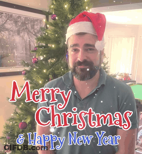 A Man Wearing A Santa Hat Stands In Front Of A Christmas Tree GIF