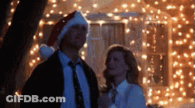 A Man Wearing A Santa Hat Stands Next To A Woman In Front Of Christmas Lights GIF