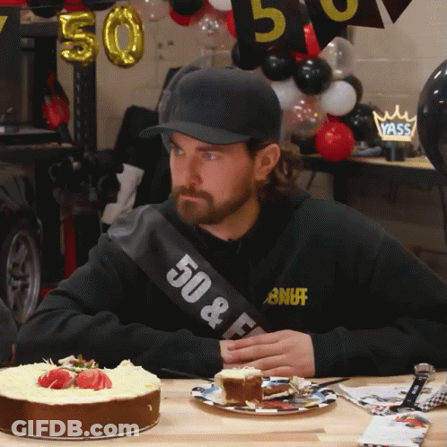 A Man Wearing A Sash That Says 50 & F Sits At A Table With A Cake GIF