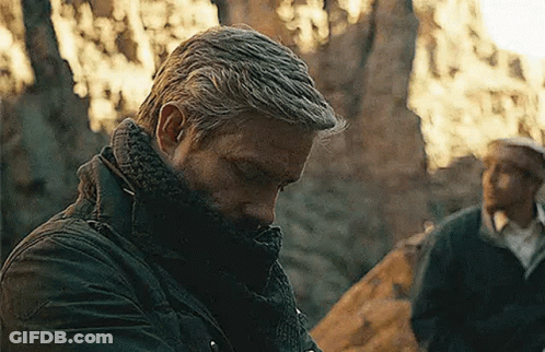 A Man Wearing A Scarf Around His Neck Is Standing In The Woods Looking Down . GIF