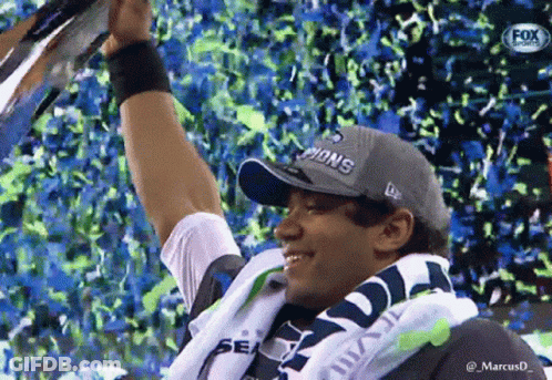 A Man Wearing A Seahawks Hat Is Holding Up A Trophy GIF