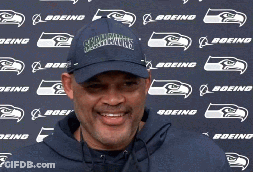 A Man Wearing A Seahawks Hat Stands In Front Of A Boeing Logo GIF