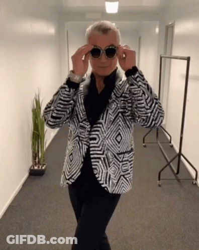 A Man Wearing A Sequined Jacket And Sunglasses Stands In A Hallway GIF