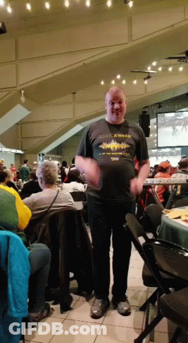 A Man Wearing A Shirt That Says X Walks Is Standing In A Crowded Room GIF