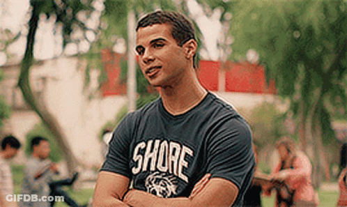 A Man Wearing A Shore T-Shirt Is Standing In A Park With His Arms Crossed GIF