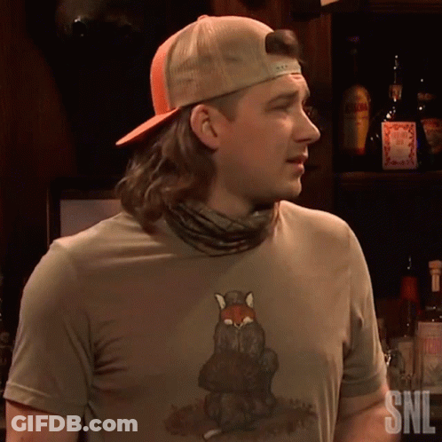 A Man Wearing A Snl Shirt Stands In Front Of A Shelf Full Of Liquor Bottles GIF