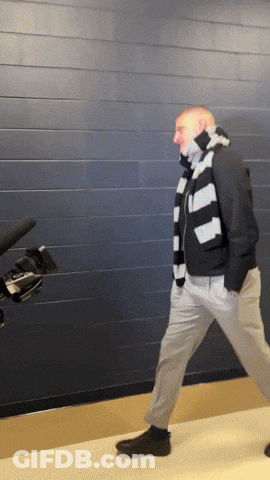 A Man Wearing A Striped Scarf And A Black Jacket Is Walking In Front Of A Camera GIF