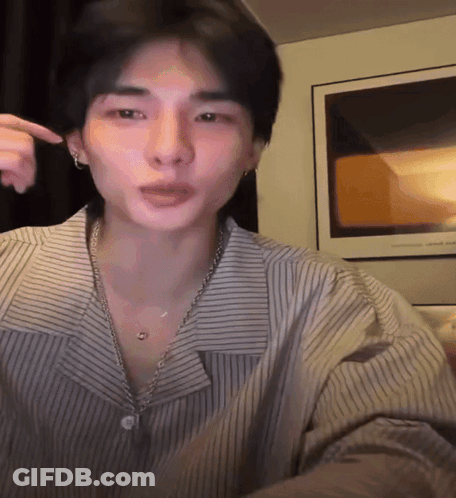 A Man Wearing A Striped Shirt And A Necklace Is Pointing At His Face GIF
