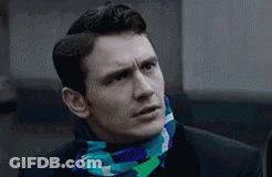 A Man Wearing A Suit And A Scarf Around His Neck Is Looking At The Camera . GIF