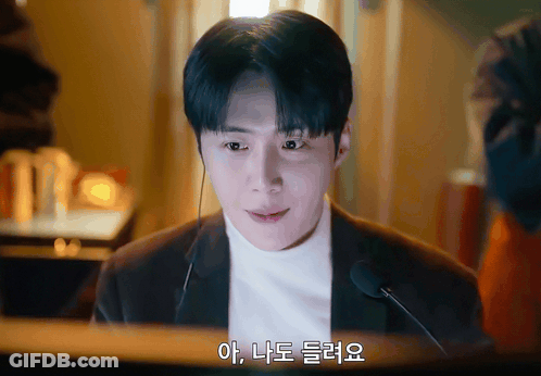 A Man Wearing A Suit And A White Turtleneck Talks Into A Microphone With Korean Writing On The Bottom Right GIF