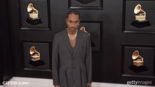 A Man Wearing A Suit And Pearls Stands In Front Of A Wall Of Grammophones GIF