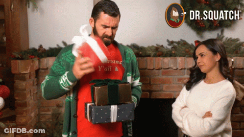 A Man Wearing A Sweater That Says Christmas Y'all Is Holding A Stack Of Gifts GIF