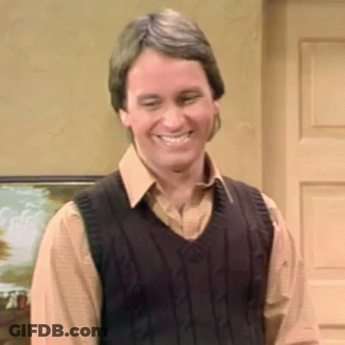 A Man Wearing A Sweater Vest And A Brown Shirt Is Smiling While Standing In Front Of A Door . GIF