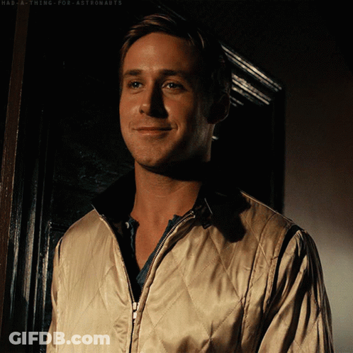 A Man Wearing A Tan Quilted Jacket Is Smiling In Front Of A Black Door GIF