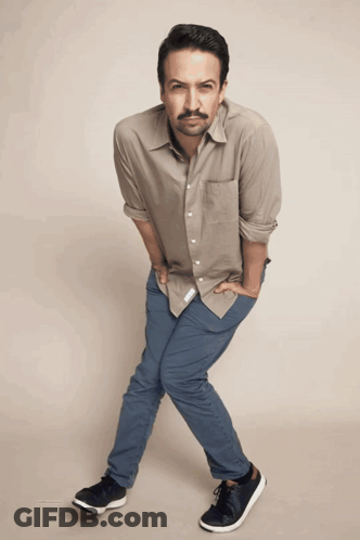 A Man Wearing A Tan Shirt And Blue Pants Is Standing With His Hands In His Pockets GIF