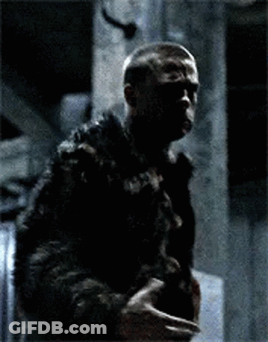 A Man Wearing A Tank Top And A Leather Jacket Is Sitting Down With His Arms Outstretched In A Dark Room . GIF