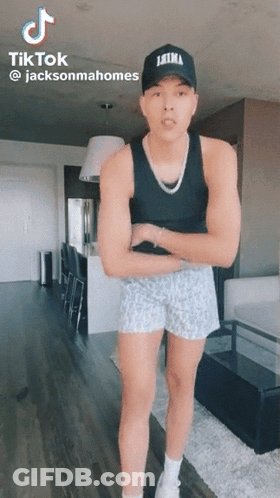 A Man Wearing A Tank Top And Shorts Is Standing In A Living Room With His Arms Crossed . GIF